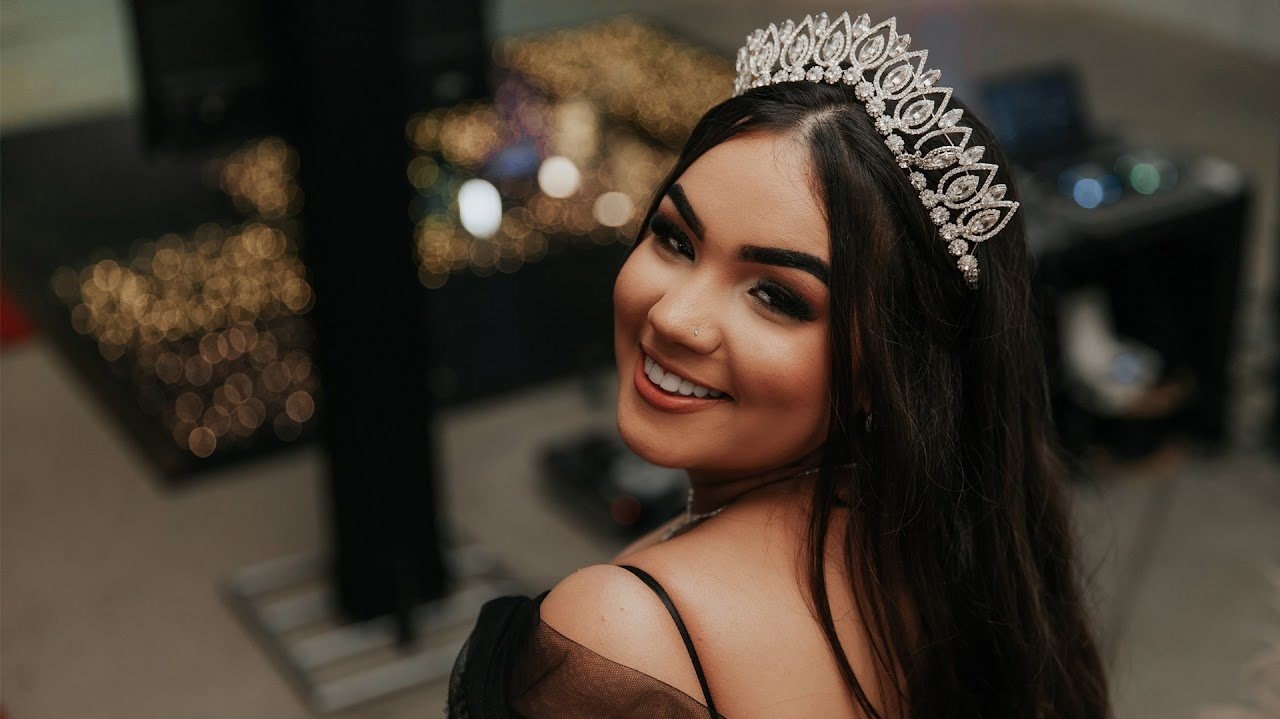 Debutante sorrindo, de tiara, com luzes desfocadas ao fundo
